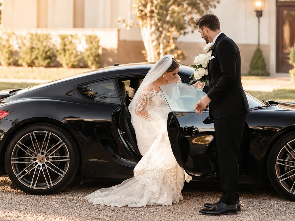 Le marié tient la portière d'une voiture sportive noire à la mariée